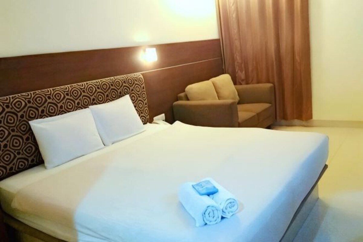 Habitación de hotel con una cama grande, cabecera con estampado geométrico, un sillón pequeño y una cortina.