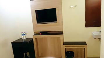 A hotel room with a bed, a TV mounted on a wooden shelf, a small desk with a chair, a bedside table, and a trash bin.