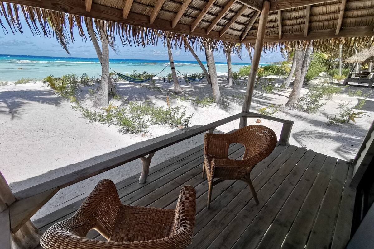 A wooden deck with wicker chairs overlooking a beach with a hammock and palm trees.