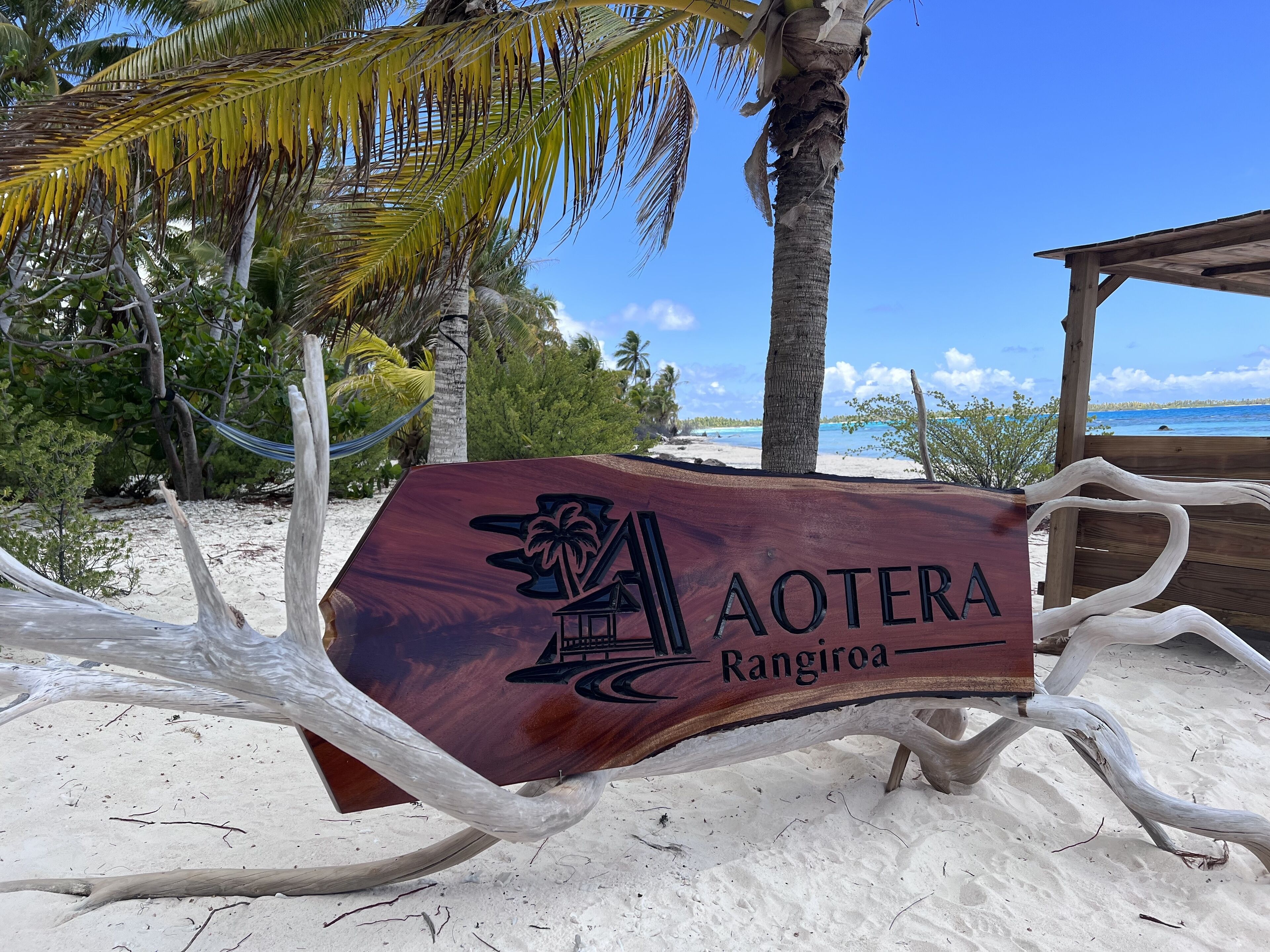 A wooden sign with 'AOTERA Rangiroa' engraved on it, surrounded by a beach setting with palm trees and a wooden structure.