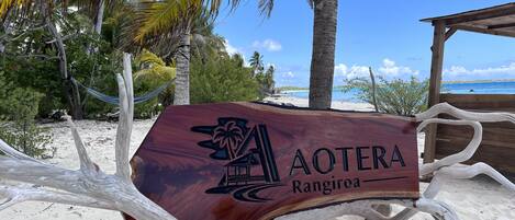 A wooden sign with 'AOTERA Rangiroa' engraved on it, surrounded by a beach setting with palm trees and a wooden structure.