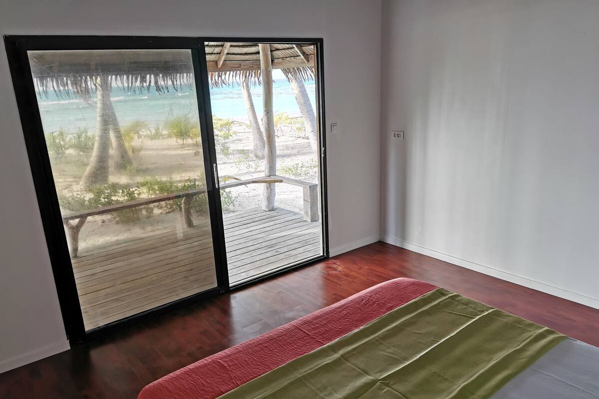 A room with a large sliding glass door leading to a wooden deck with a view of the beach and ocean.
