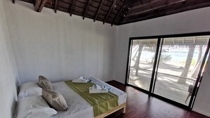 A bedroom with a wooden bed, a green blanket, and a view of a beach through large windows. - Aotera Guest House (Rangiroa)