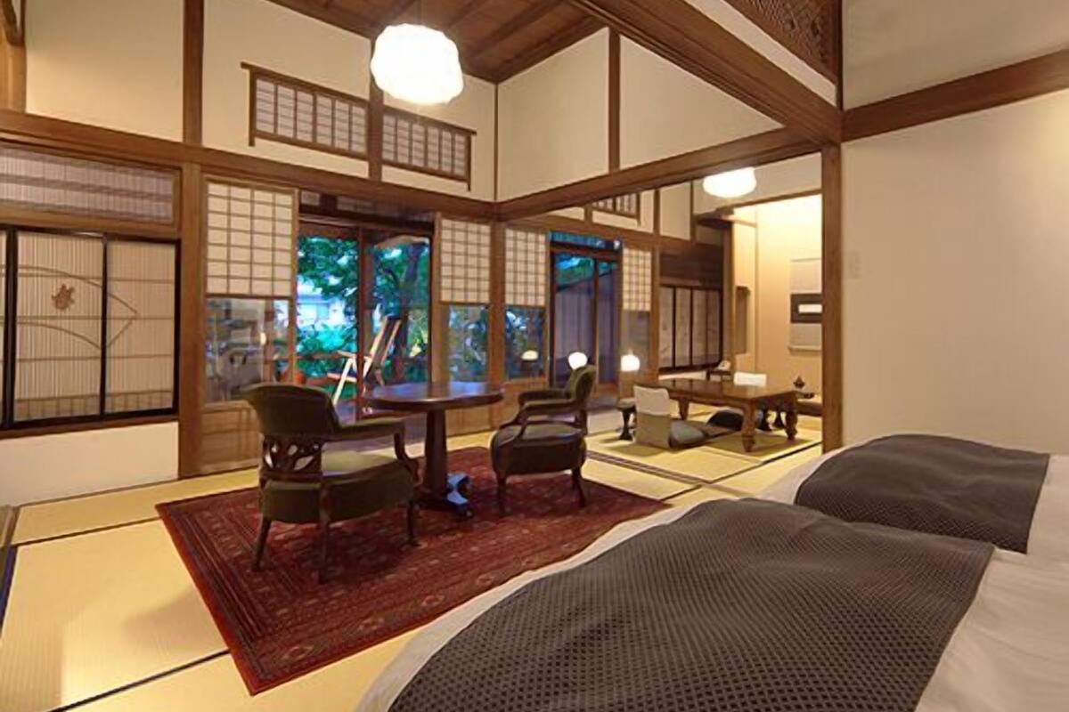Une pièce de style japonais traditionnel, avec un sol recouvert de tatamis, une table basse, des chaises et un lit recouvert d’une couverture sombre.