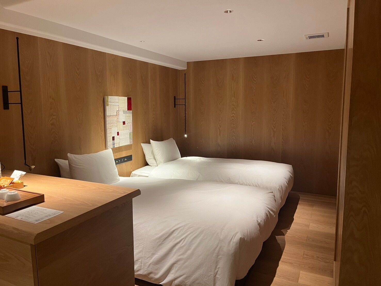 A hotel room with a wooden wall panel, a bed with white bedding, a desk with a lamp, and a bulletin board.