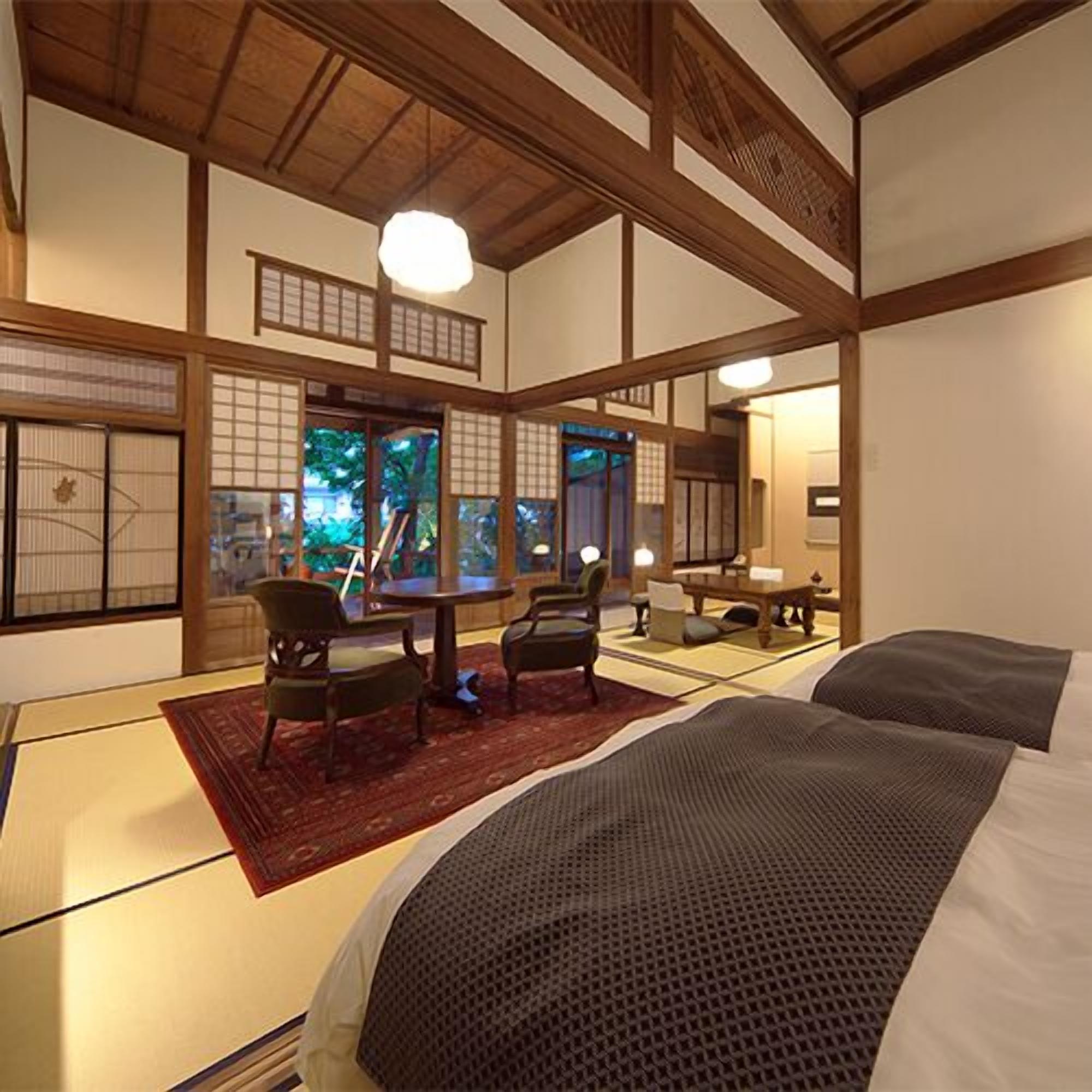 A traditional Japanese-style room with tatami flooring, a low table, chairs, and a bed with a dark blanket.