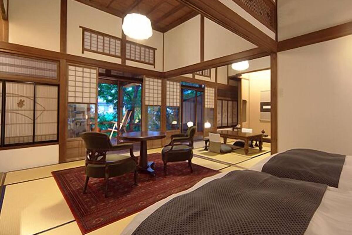 Une pièce de style japonais traditionnel, avec un sol recouvert de tatamis, une table basse, des chaises et un lit recouvert d’une couverture sombre.