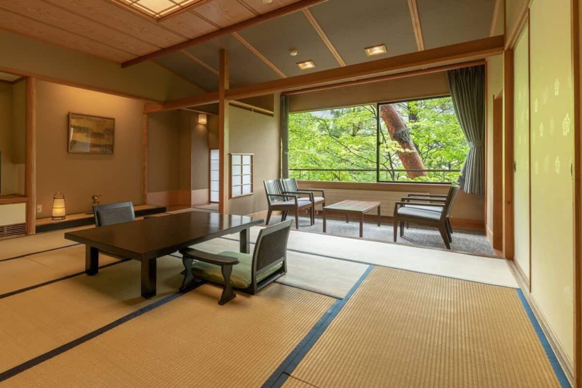 Una habitación tradicional japonesa con suelo de tatami, una mesa baja, sillas, un televisor y un gran ventanal con vistas a árboles.