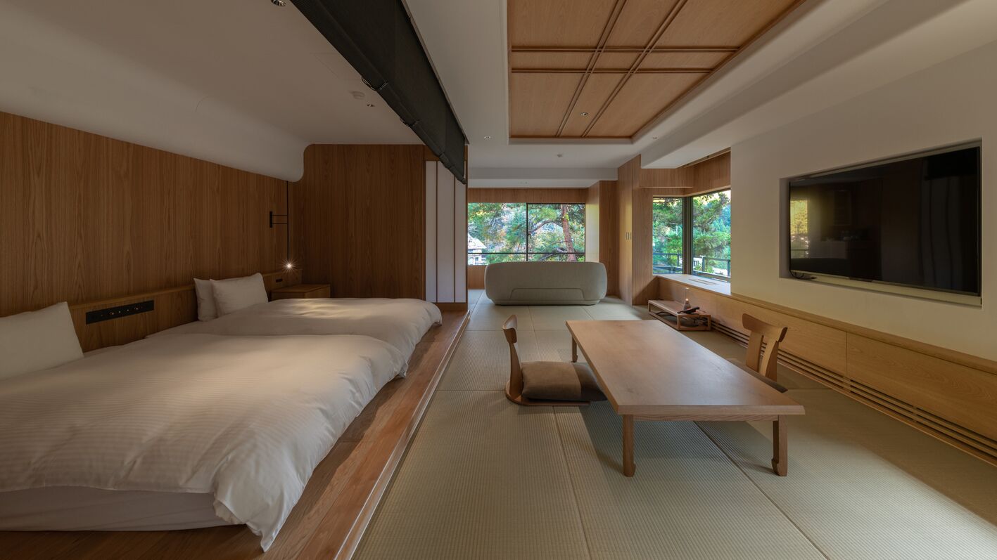 A modern hotel room with two beds, a dining table, and a large window offering a view of trees.