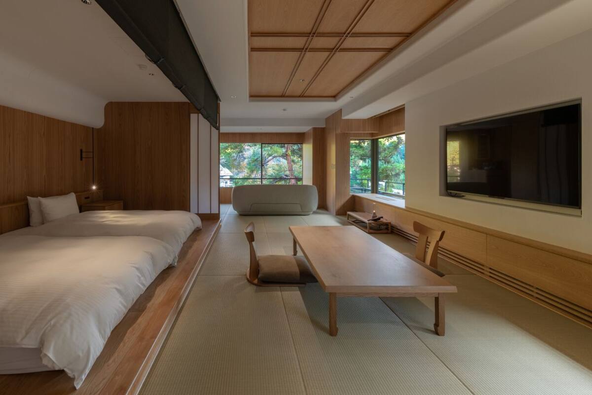 Une chambre d’hôtel moderne avec deux lits, une table à manger et une grande fenêtre donnant sur un paysage arboré.