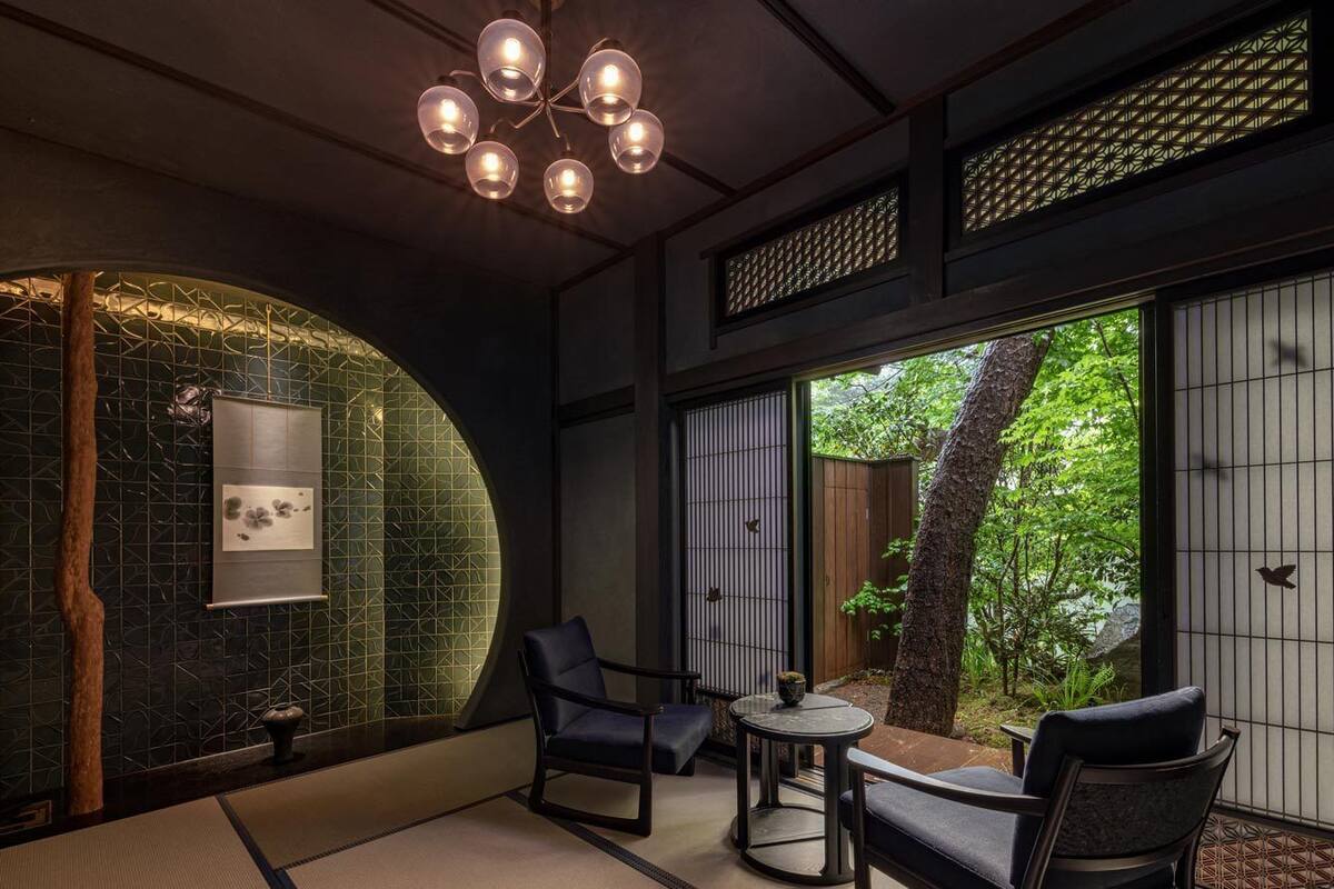 Une pièce de style japonais traditionnel, avec un sol recouvert de tatamis, une table basse entourée de chaises, un luminaire suspendu et une vue sur la verdure à l’ext érieur.