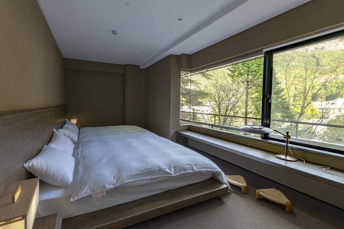 Une chambre à coucher moderne avec un grand lit, une fenêtre donnant sur la verdure et une petite table de chevet.