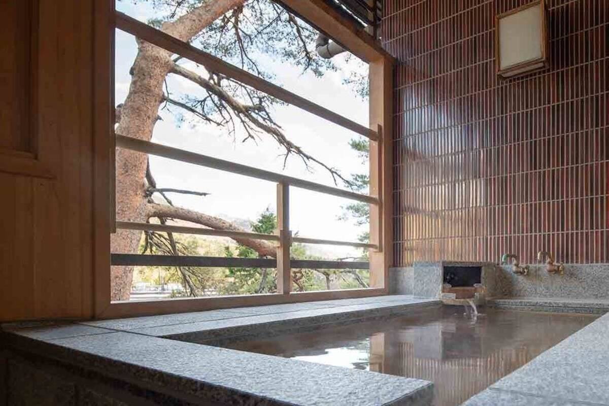 Un onsen japonais traditionnel, avec un bain en pierre, des portes coulissantes en bois et une vue sur un arbre à l’extérieur.
