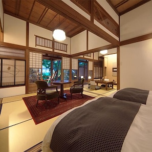 Una habitación de estilo japonés tradicional con suelo de tatami, una mesa baja con sillas, una cama con una manta oscura y puertas correderas que dan a una vista de follaje.