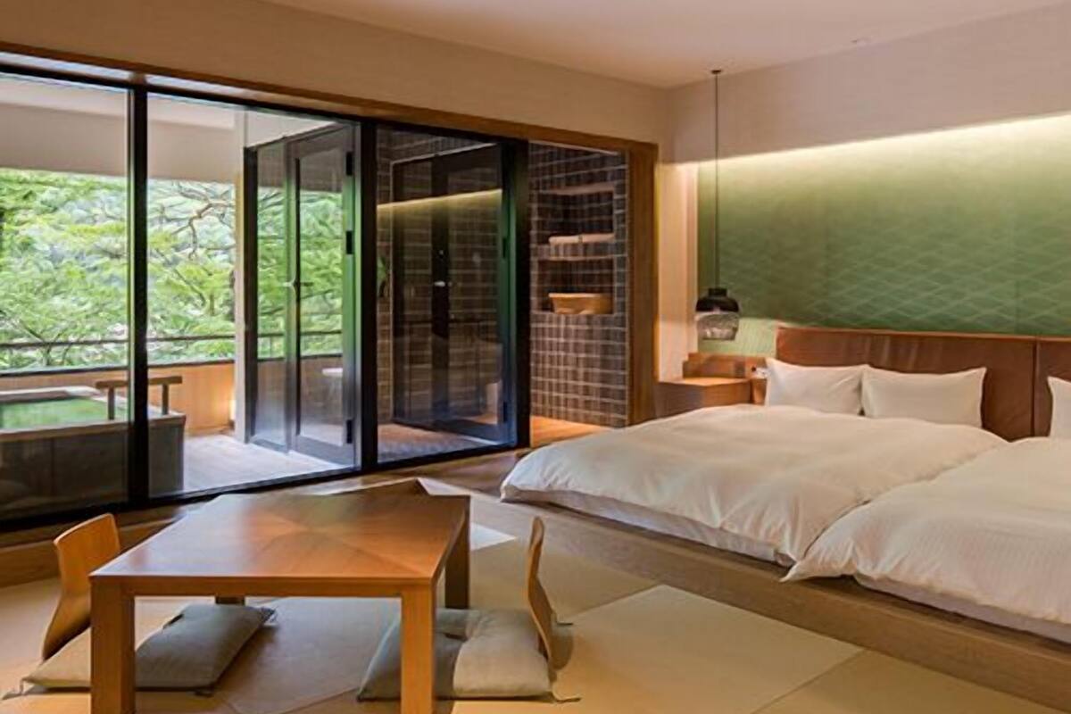 Une chambre d’hôtel moderne dotée d’une grande porte-fenêtre coulissante donnant sur un balcon, d’une table en bois et d’un lit avec du linge de lit blanc.