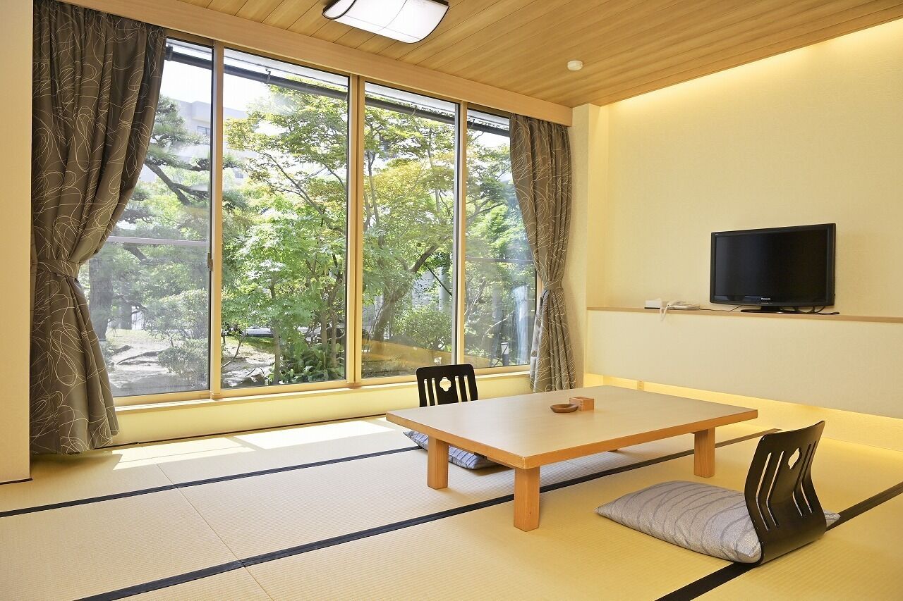 A room with tatami flooring, a wooden table, chairs, a television on the wall, and large windows with curtains.