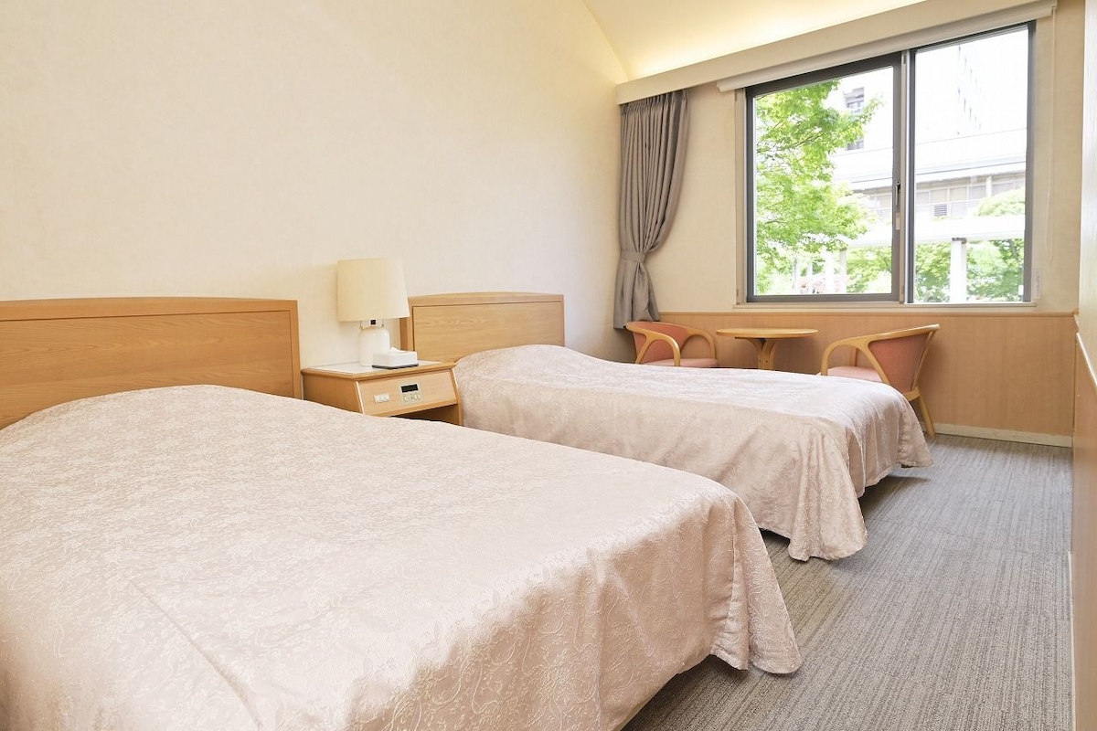 Une chambre d’hôtel avec deux lits, une table de chevet avec une lampe, une fenêtre avec des rideaux et une vue sur les bâtiments environnants.