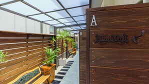 A wooden welcome sign with a metal 'A' and 'B' indicating rooms, a bench, potted plants, and a tiled pathway. - Orion Resort (Tokyo)