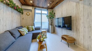 A modern living room with a grey sofa, a wooden coffee table with flowers, a wicker chair, a flat-screen TV, and a large window with curtains. - Orion Resort (Tokyo)