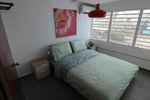 A bedroom with a bed, a nightstand, a small table, a window with blinds, and a wall with a painting.