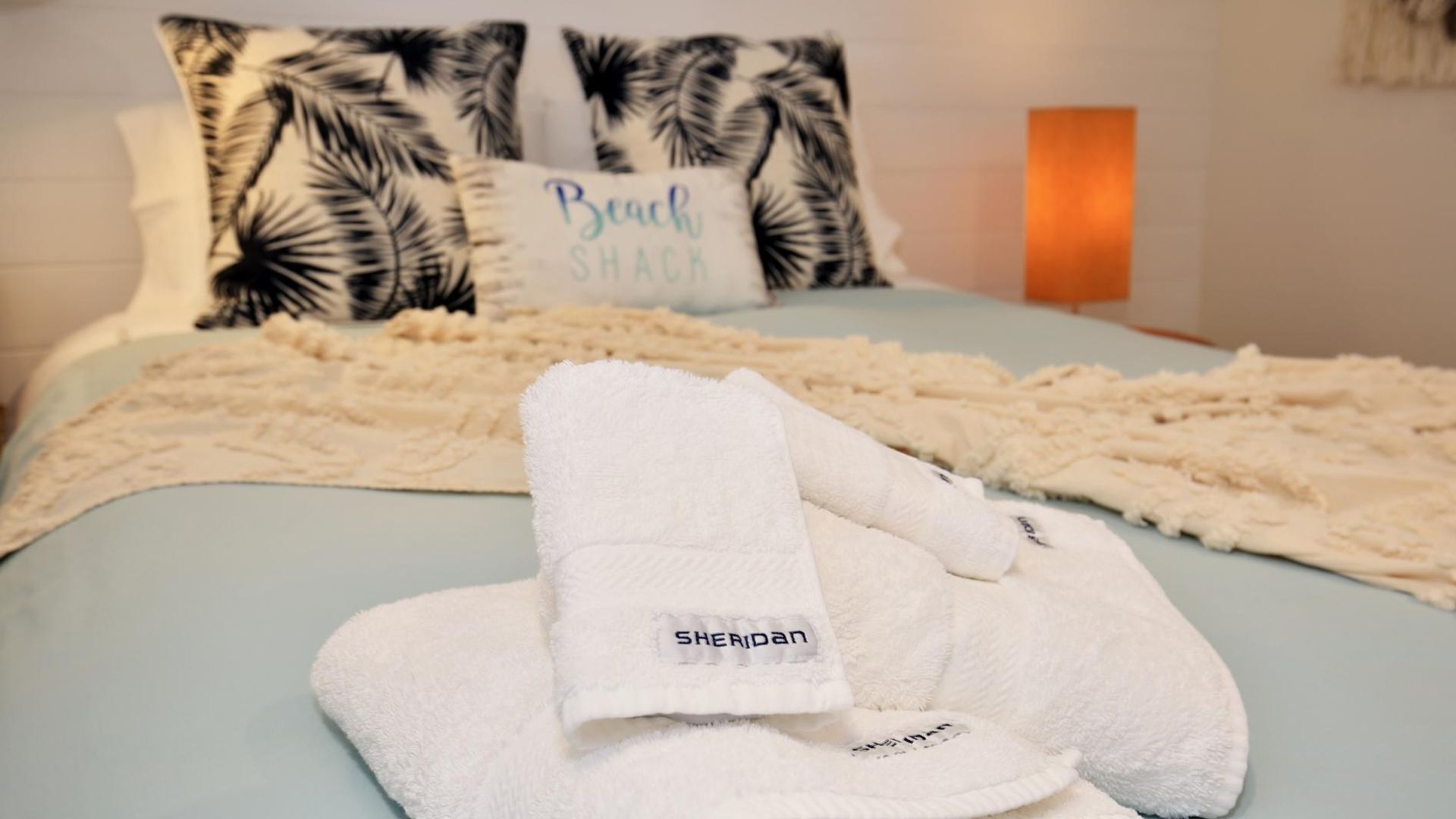 A bed with a white blanket, a white towel, and a pillow with 'Beach Shack' text.