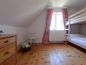 Ein Zimmer mit Holzboden, einem Etagenbett, einem weißen Heizkörper, einem Fenster mit roten Karottenvorhängen, einem weißen Stuhl und einer Holzkommode mit Schubladen.