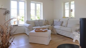 Un salon spacieux avec un grand canap é blanc, un petit pouf et une table noire. Deux fenêtres laissent entrer la lumière naturelle, et un radiateur noir est visible sur le côté droit.