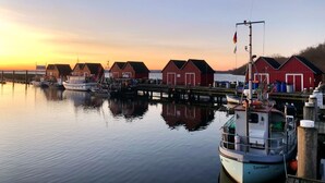A harbor with several red wooden buildings, boats docked, and calm water reflecting the structures. - Urlauberdorf 40b, Deutschland (Ostseebad Boltenhagen)