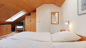 A bedroom with a sloped ceiling, wooden walls, a skylight, a bed with white linens, a chair, and a bedside table with a lamp. - Urlauberdorf 40b, Deutschland (Ostseebad Boltenhagen)