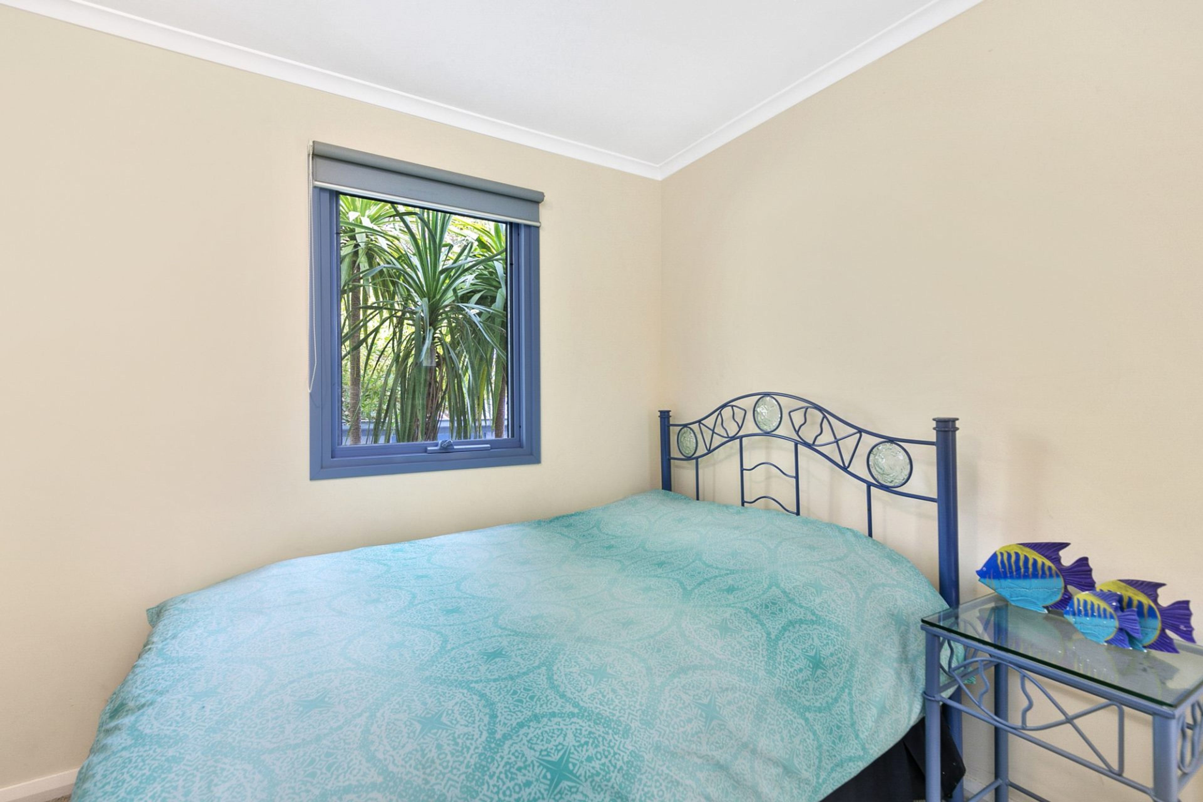 A bedroom with a bed, a window with a view of palm trees, and a small table with a fish decoration.