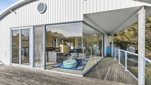 A modern building with large glass windows, a wooden deck, and a visible interior with furniture. - Caprica (Fairhaven)