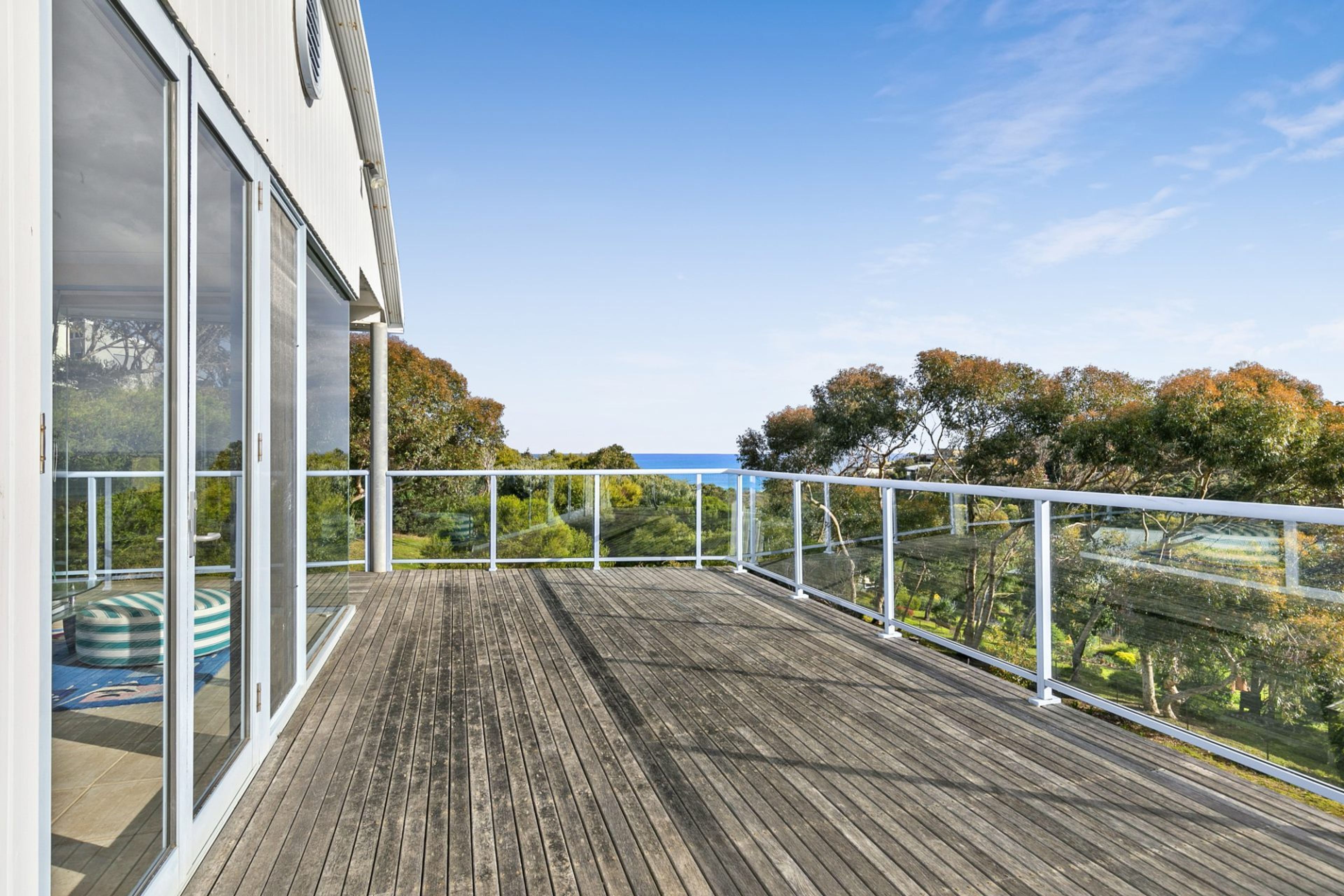 A wooden deck with glass railings overlooking a landscape with trees and a view of the sea.