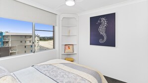 A bedroom with a bed, a window with a view of buildings, a ceiling fan, and a framed artwork on the wall. - Beachpoint Unit 503 (Forster)