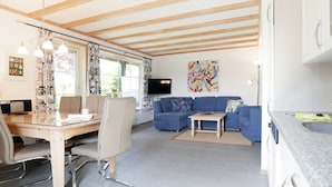 Een moderne woonkamer met een houten plafond, een blauwe bank, een eettafel met stoelen en een keuken met een aanrechtblad en spoelbak.
