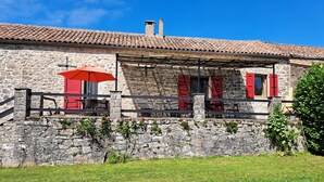 A stone building with a tiled roof, outdoor seating with red chairs and an umbrella, and a grassy lawn.