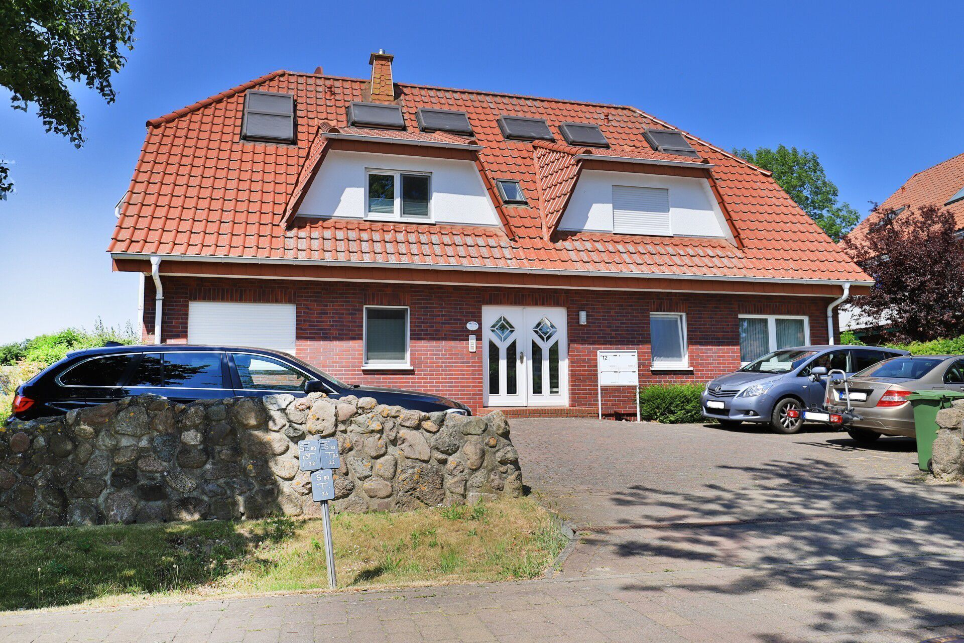 Een huis met twee verdiepingen, voorzien van een rode dakbedekking, een stenen muur ervoor en twee auto's op de oprit.