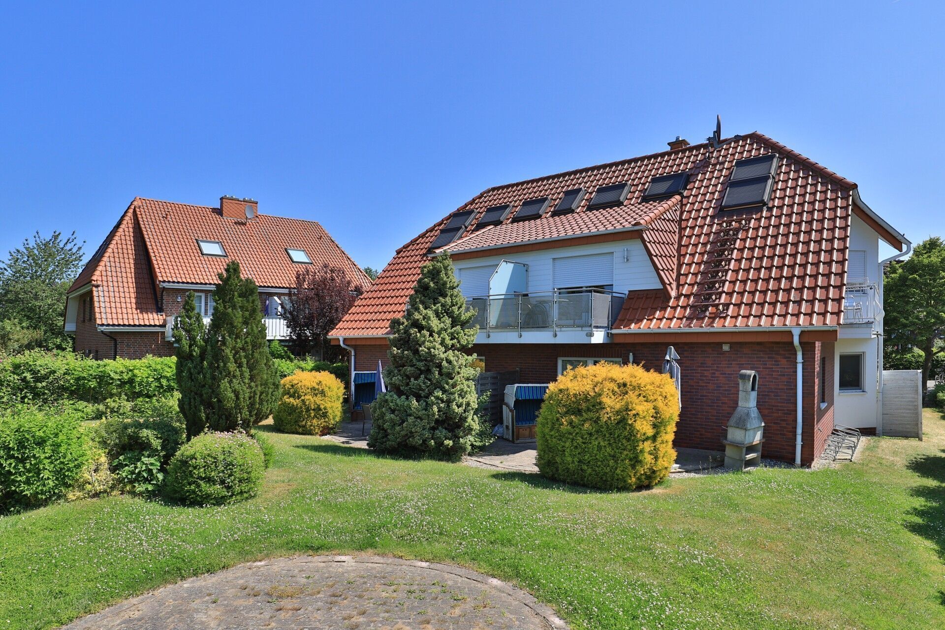 Ein zweigeschossiges Wohnhaus mit rotem Ziegeldach, einem Balkon und einem Garten mit Hecken und Rasen.
