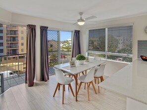 Un comedor moderno con una mesa y sillas blancas, un ventilador de techo y vistas a la ciudad a través de amplias ventanas con persianas.