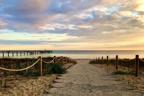 Een verhard pad dat naar een strand leidt, met een houten steiger op de achtergrond.