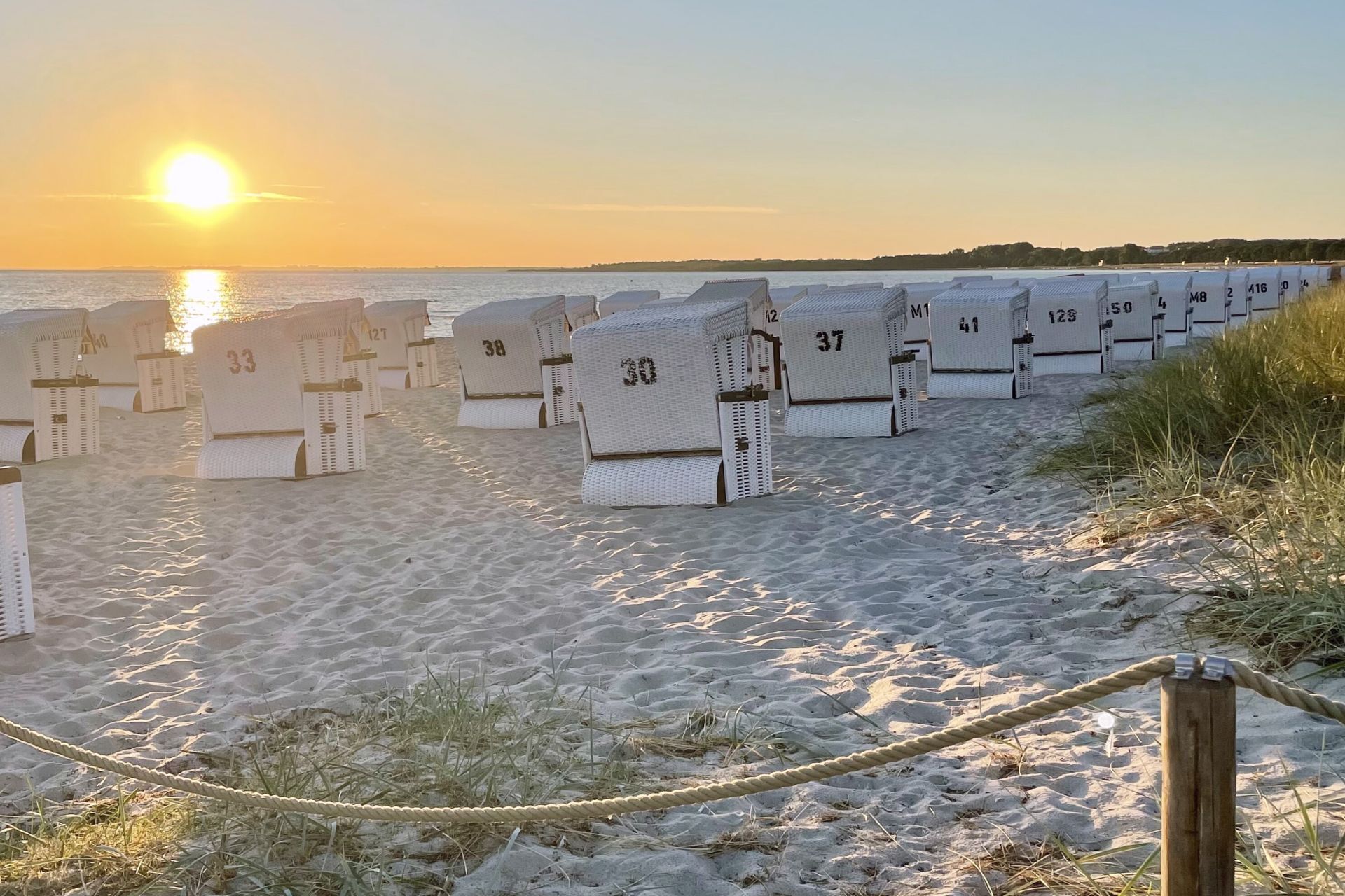 A row of numbered beach huts on a sandy beach with the sun setting in the background.