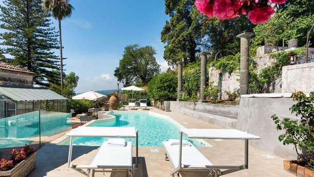 Ein Poolbereich mit Liegestühlen und Sonnenschirmen, umgeben von Bäumen und einer Steinmauer.