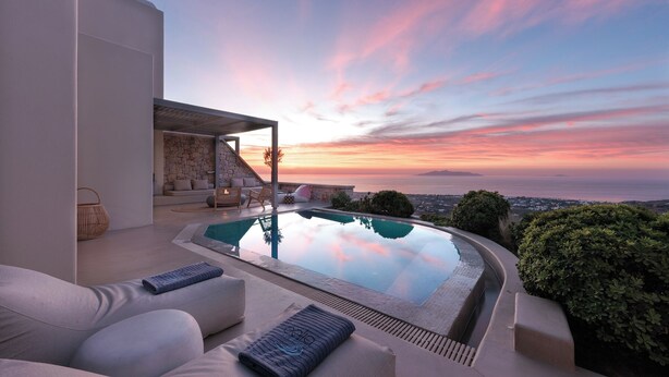 Uma área de piscina moderna ao ar livre, com muro de pedra, espreguiçadeiras e vista para o pôr do sol sobre o oceano.