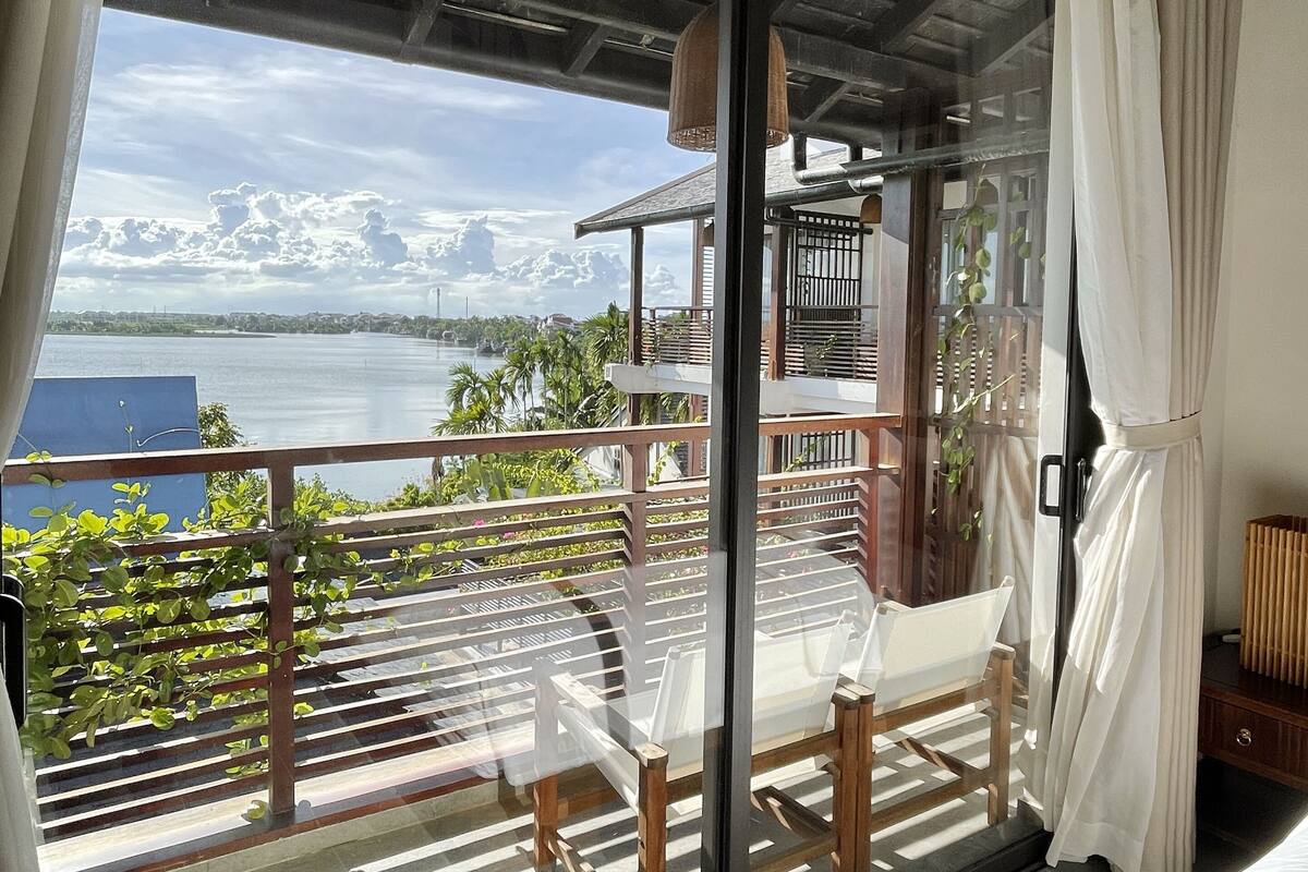 Varanda com vista para o mar e montanhas, uma cadeira de madeira, uma cama e uma pequena mesa.