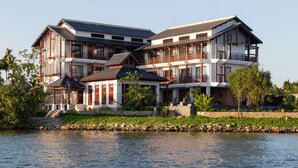 A large, multi-story building with a prominent balcony, situated by a body of water. - Nghe Prana Hotel and Spa Hoi An (Da Nang)