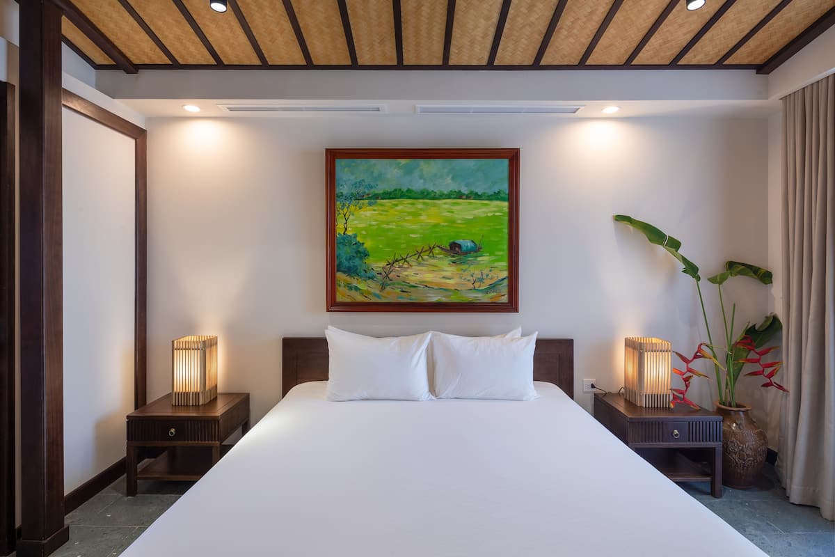 Quarto de hotel com uma cama grande, duas mesas de cabeceira com abajures, um quadro na parede e um vaso com planta.