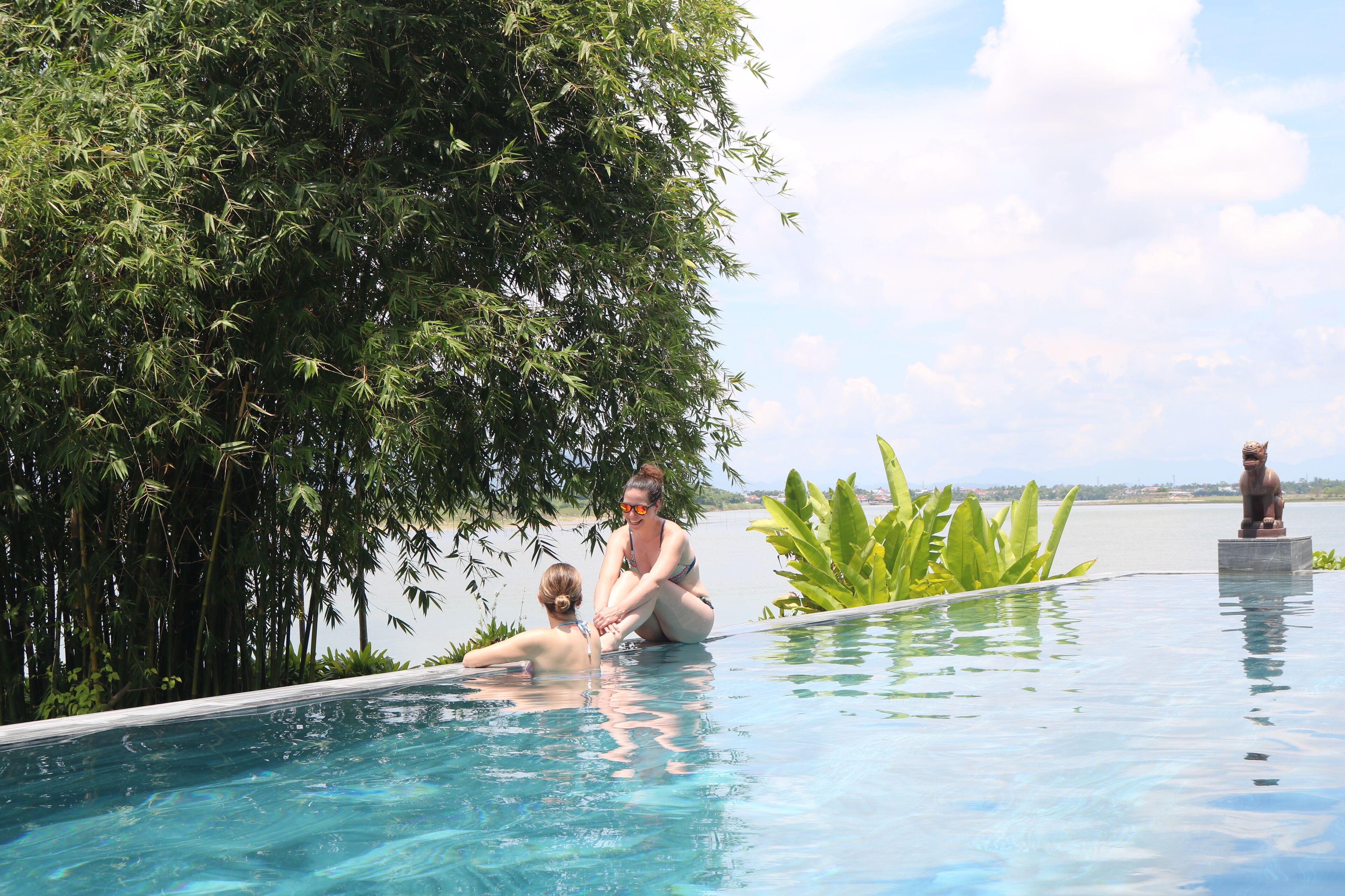 A pool with two people sitting on the edge, surrounded by lush greenery and a statue.
