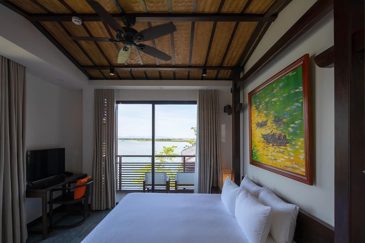 Quarto de hotel com cama, escrivaninha com televisão, pintura na parede e vista para a água.