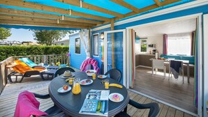 Een overdekt terras met een eettafel voor twee personen, een klein keukenhoekje en een blauw huis op de achtergrond.