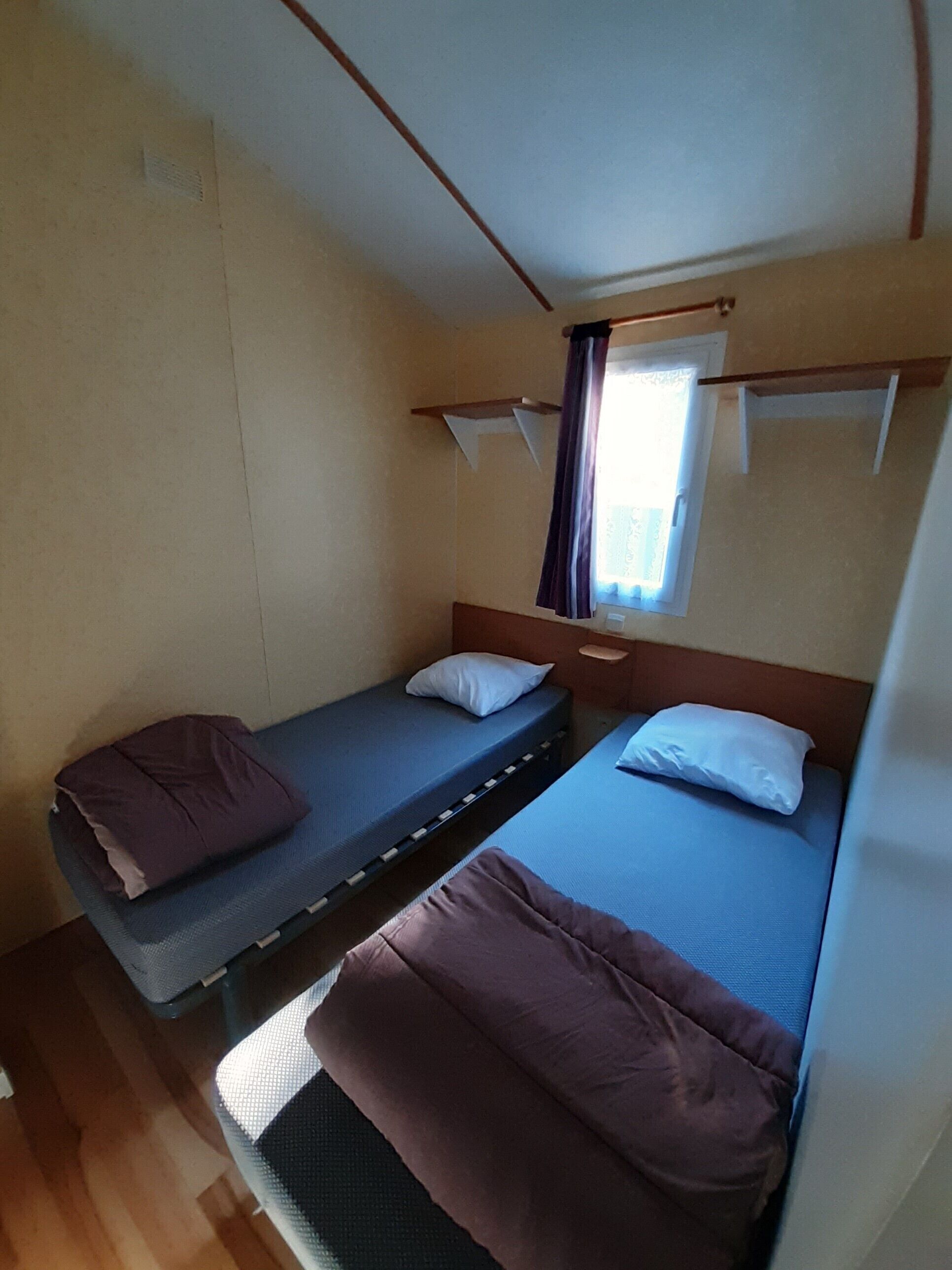 A compact room with two single beds, a small window with curtains, and a wooden headboard.