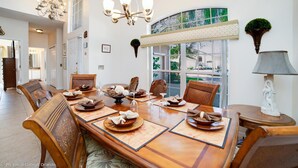 Une salle à manger avec une grande table en bois, dressée pour six personnes, entourée de chaises en bois. Un lustre est suspendu au-dessus de la table et une fenêtre offre une vue sur l’extérieur.